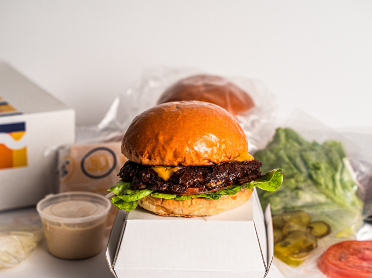 A burger sits atop a white box, vac packed ingredients in background