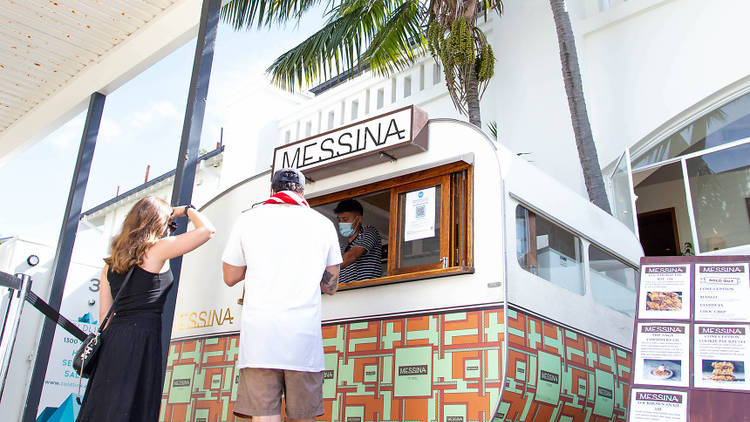 Two people queue up at the service window of the Messina caravan pop-up outside the Harbord Hotel