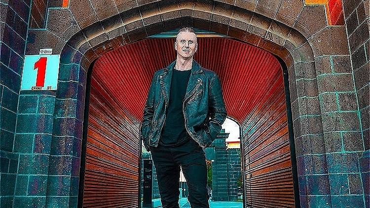 A man wearing a denim jacket and black boots stands outside Pentridge Prison. He has one foot on a skateboard that's covered in paint