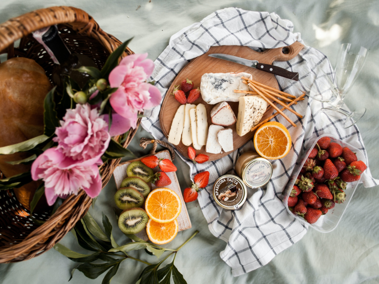 A flat lay of a picnic with cheeses, fresh fruit, a gingham rug and pink flowers