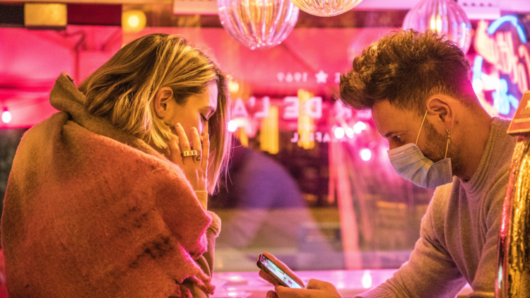 Two people on a date, a man wears a mask and looks at his phone while a woman looks over.