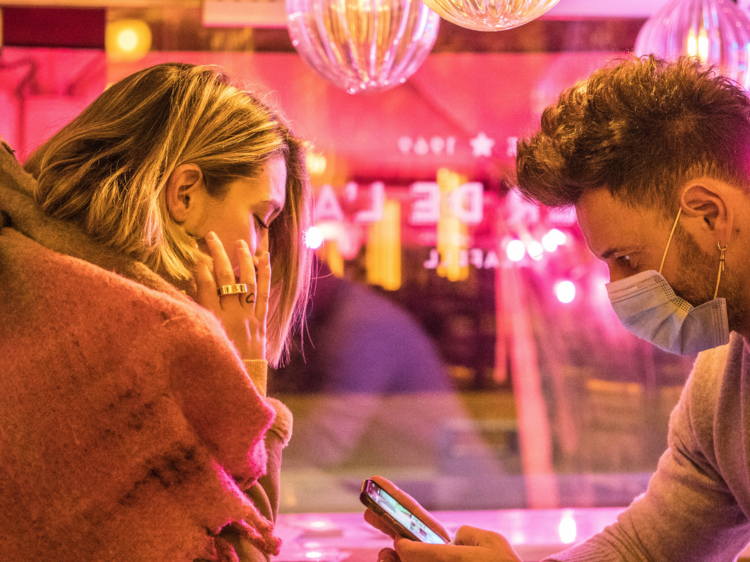 Two people on a date, a man wears a mask and looks at his phone while a woman looks over.