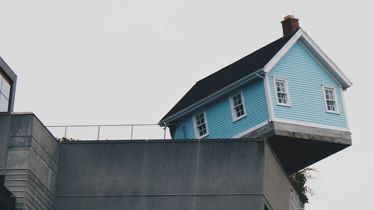 A pale blue-painted weatherboard house wobbling precariously on the edge of another building