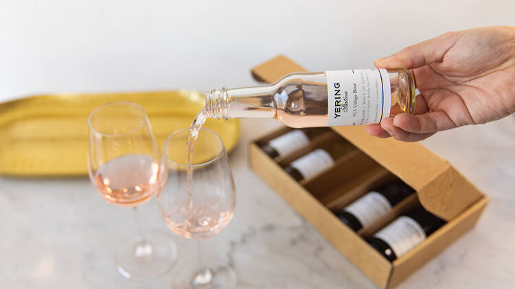 A small bottle of rosé wine is poured into two wine glasses, with a box of more wines in the background