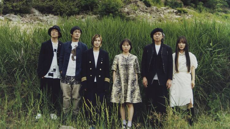 Six members of the band Leenalchi stand shoulder-to-shoulder in front of tall green grass
