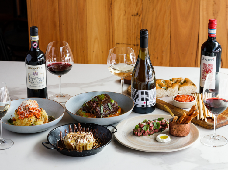 A table full of bowls of food, steaks, potatoes, bottles of wine, wine glasses