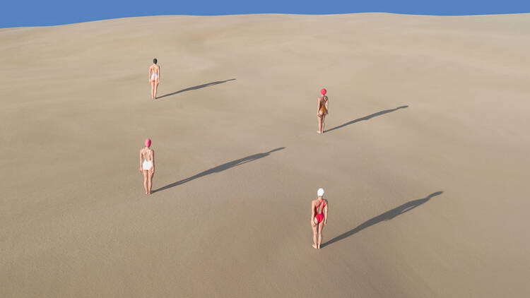 Three women in swimsuits standing on a sand dune casting shadows with blue sky above