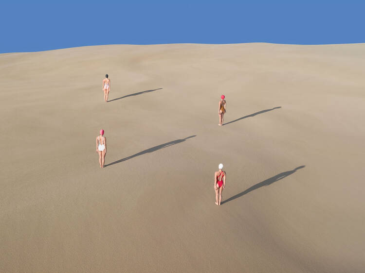 Three women in swimsuits standing on a sand dune casting shadows with blue sky above