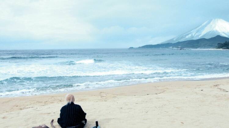 A man sits on a beach with crashing wave and a snow capped mountain in the distance