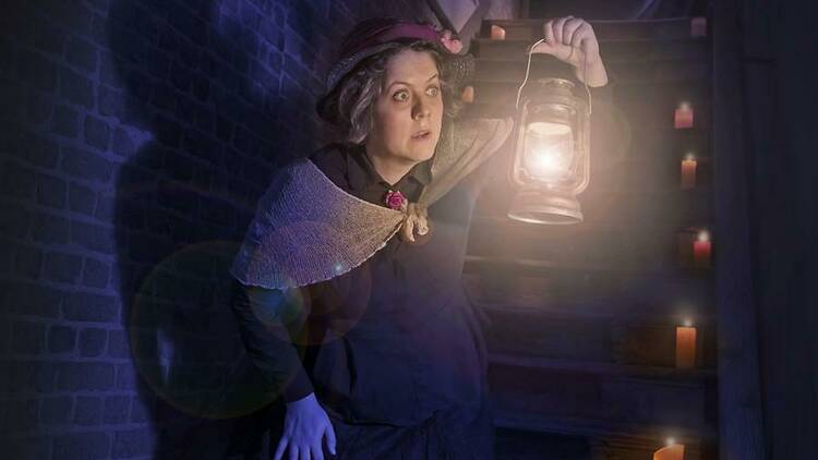 A scared looking woman dressed in old fashioned clothing holds a lantern