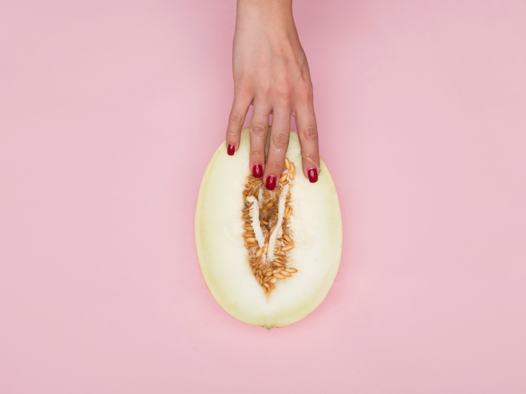 A hand with fuchsia nail polish delicately touches a cut piece of fruit that has an abstract resemblance to a vulva