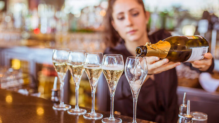 A woman pours a bottle of champagne into five glasses lined up on a bar