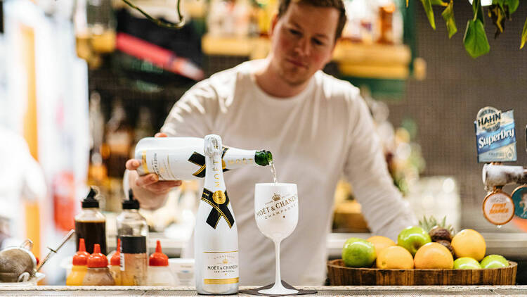 A man in a white long-sleeve top pours a bottle of champagne into a white, Moet & Chandon branded glass