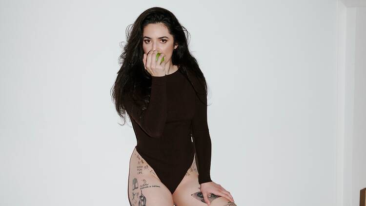 Musician Georgia Maq wears a long-sleeve leotard as she leans against a stark white wall and bites into an apple