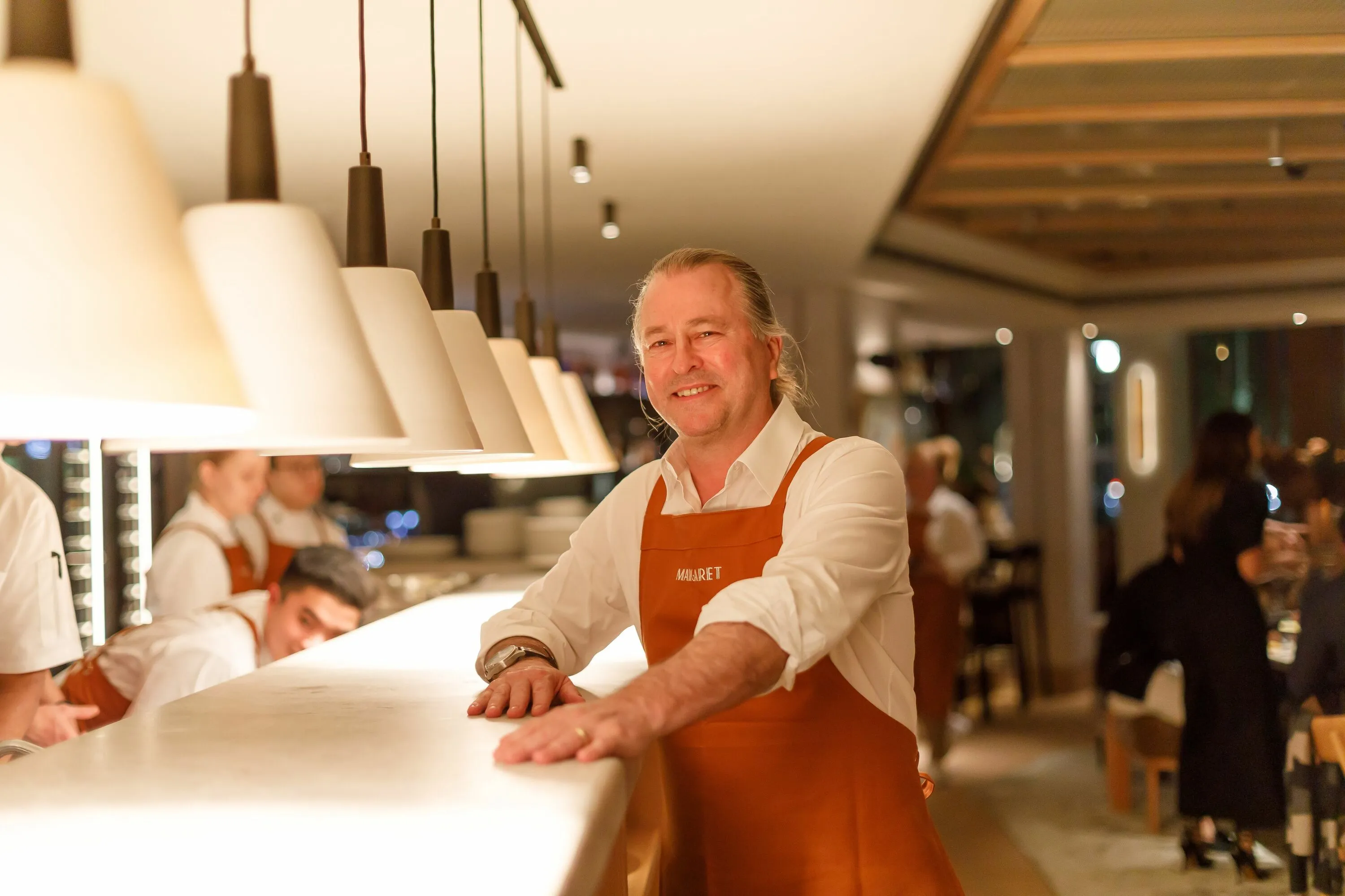 Celebrated chef Neil Perry stood next to a kitchen bench wearing a terracotta apron with the world Margaret on the front
