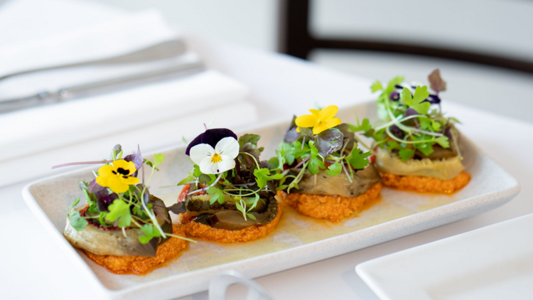 A square plate with four canapes topped with edible flowers sits on a white table cloth
