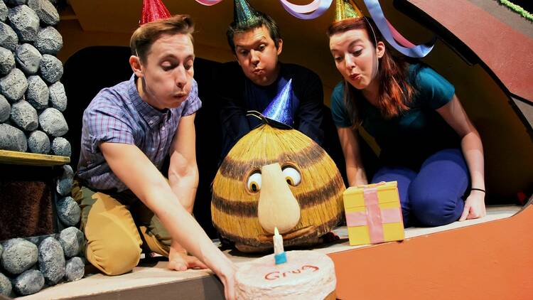 a puppet of childrens book character Grug with three humans holding a birthday cake with candle for him