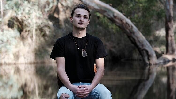 A young man sits with his hands in his lap on a chair in front of a creek in the bush