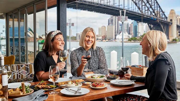 Three woman sit at table with food and wine, the Sydney Harbour Bridge is in the background
