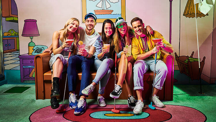 Five friends holding golf clubs and colourful cocktails grin and embrace as they sit on the famous couch tableau from The Simpsons.