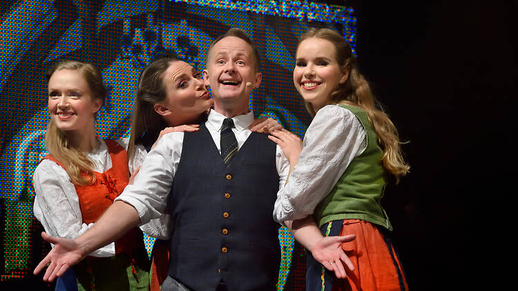 Actor Billy Boyd stands cheerfully with arms outstretched while three women wearing hobbit-style, dirndl-like dresses gather playfully around him