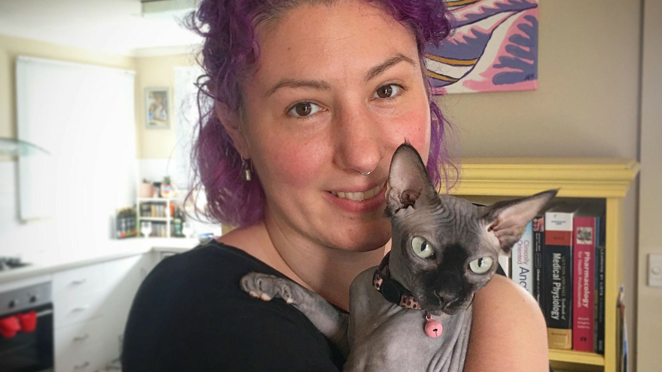 A woman with purple hair holds a hairless cat in front of a bookcase