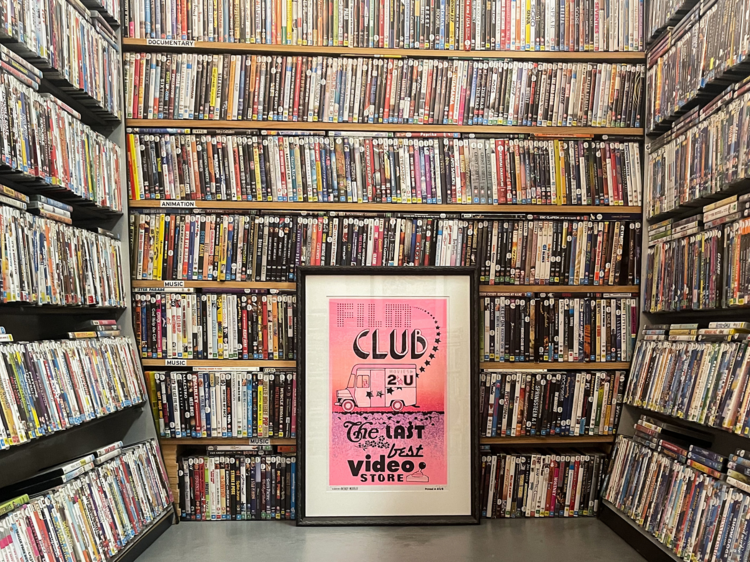 A pink poster for Flim Club: the last and best video store sits on the floor amongst shelves of DVD cases