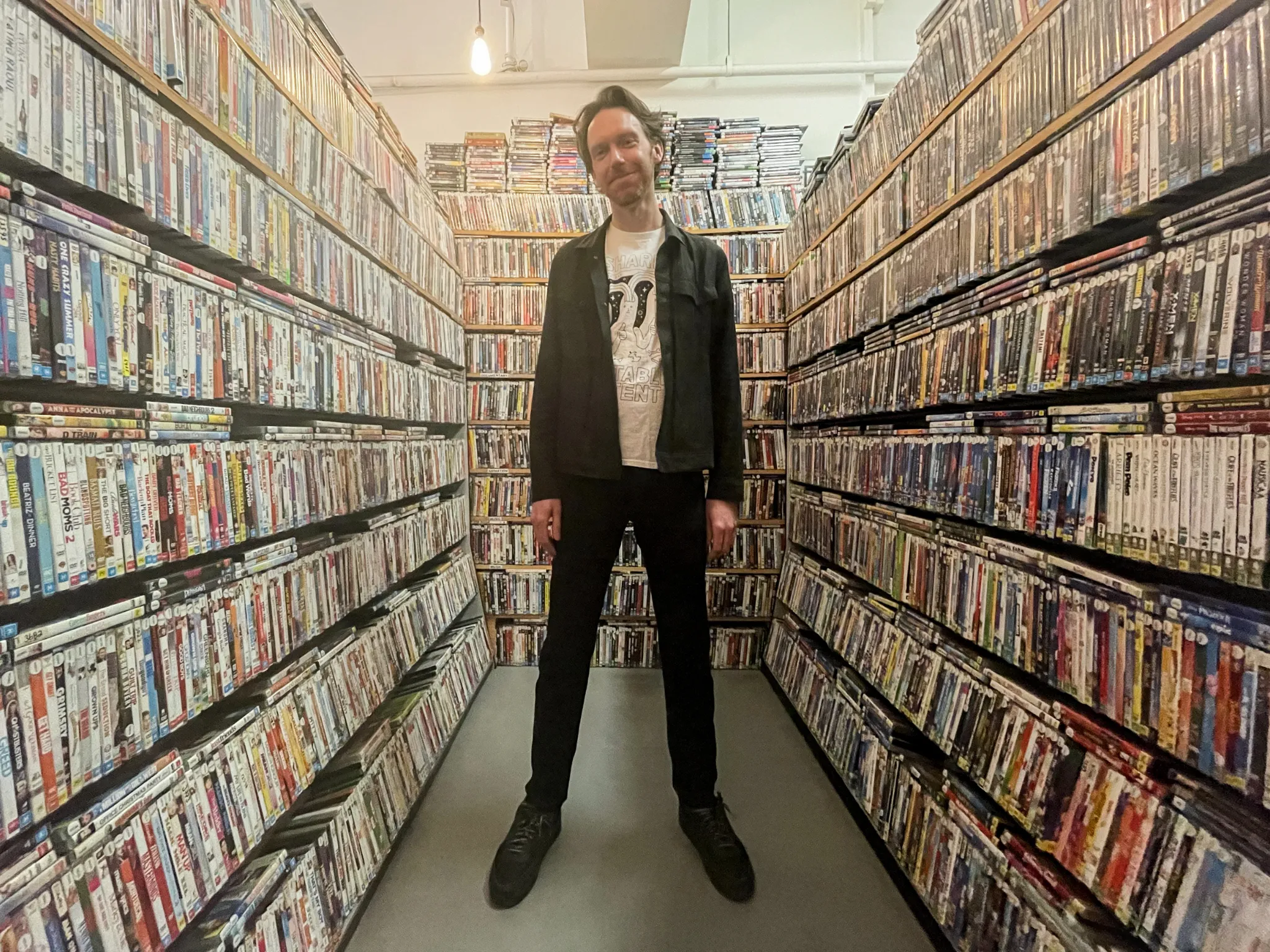 Film Club owner Ben Kenny stands amongst shelves stacked with DVD cases. He wears black skinny jeans a black jacket and a white printed t-shirt.