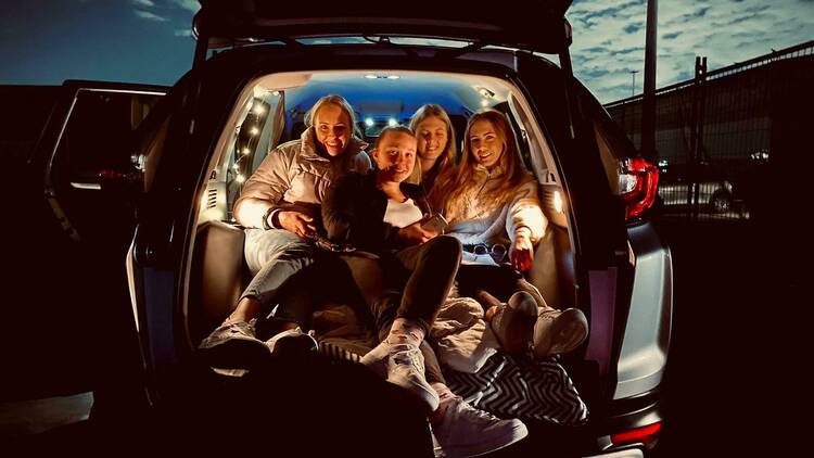A group of friends are packed into the back of a van at a drive-in cinema with the boot open, ready to watch a movie