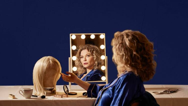 Leeanna Walsman in a brilliant blue jacket looking at us reflected in a dressing room mirror framed by lightbulbs