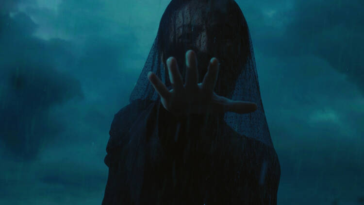 A dark veiled woman in funeral garb reaches out menacingly with a stormy sky behind her