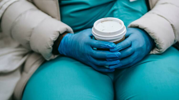 A healthcare worker wearing gloves and scrubs holds a takeaway coffee cup