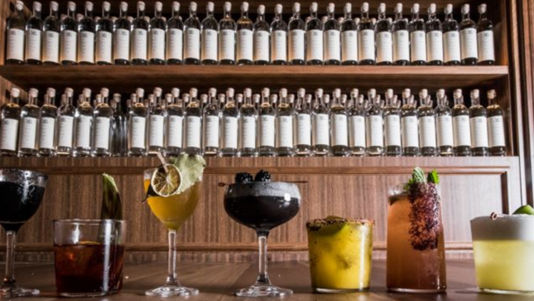 Seven cocktails lined up on a wooden bench in front a stocked bar