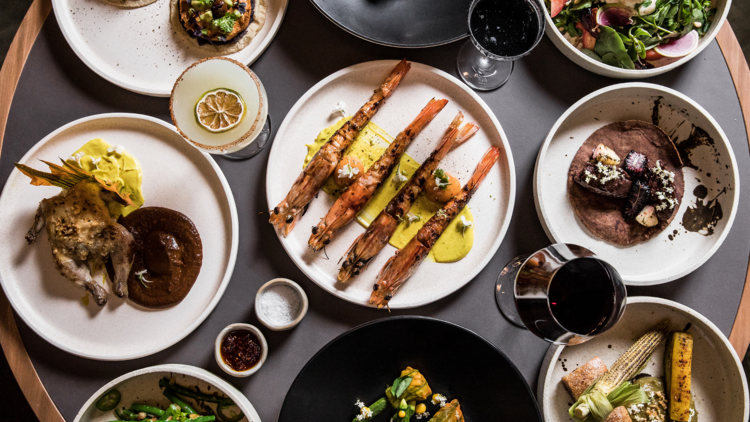 A flat lay of dishes including prawns, chicken with mole, grilled vegetables and glasses of wine