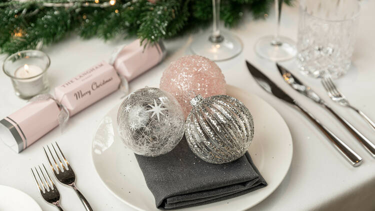 Three sparkly baubles on a plate as part of a Christmas-themed place setting. There is a pink cracker and fake pine needles with a small tealight candle setting the festive mood.