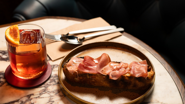 On a marble table sits a piece of bread topped with proscuitto, a negroni on the left