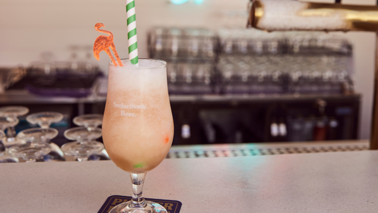 A pink shaken cocktail garnished with a flamingo on a white counter with beer taps in the background