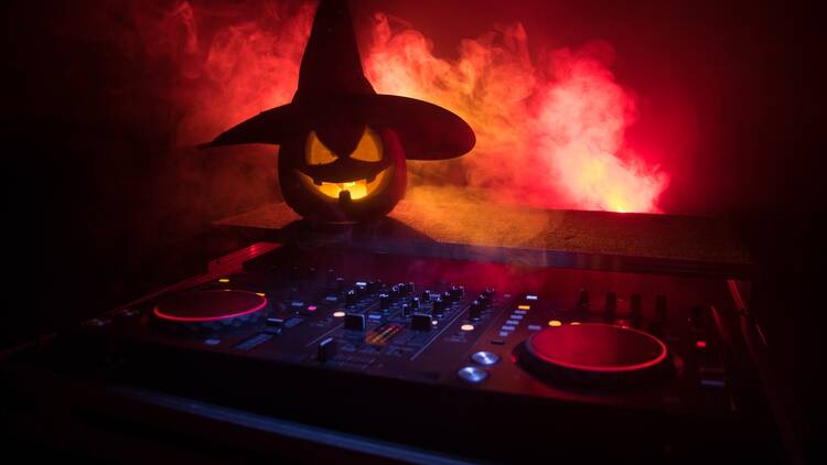 A pumpkin carved with a grinning face and wearing a witch hat, rested on some DJ decks