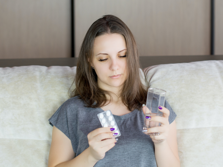 A hungover women in bed staring at a packet of panadol with a glass of water in her hand