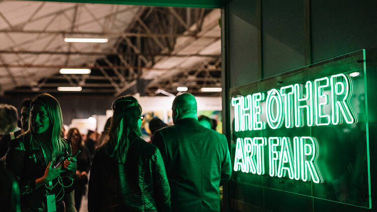 Two people walk past a green neon light that reads: The Other Art Fair