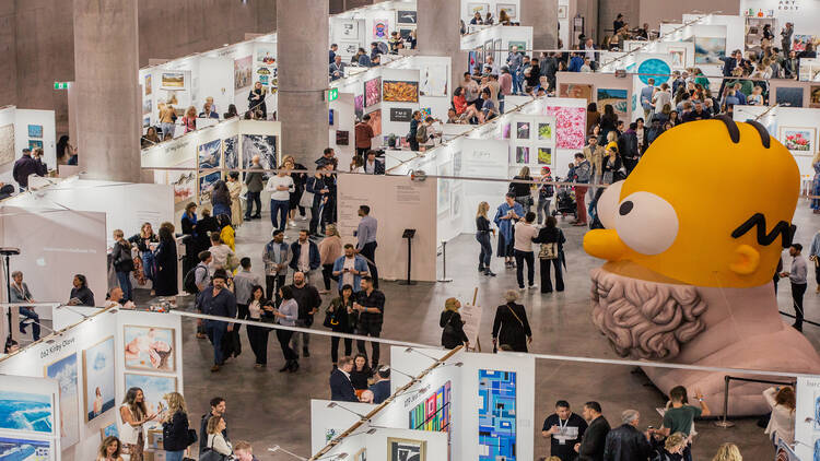 A wide shot of an art fair with crowds of people wandering around and lots of art on walls