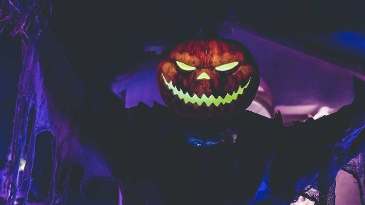 A dark figure with a glowing pumpkin head stands against a purple-lit background