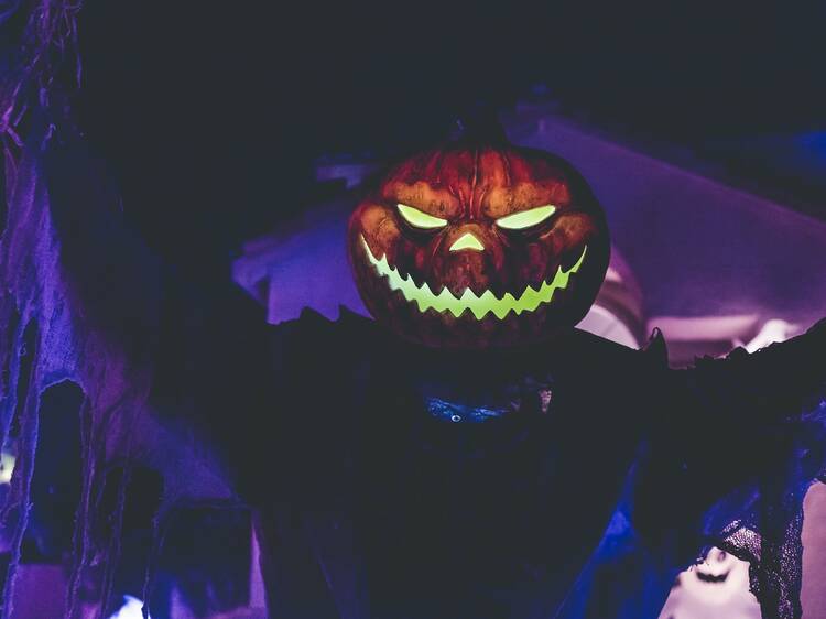 A dark figure with a glowing pumpkin head stands against a purple-lit background