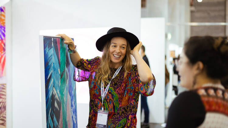 A woman wearing a black broad-brim hat holds up a piece of artwork