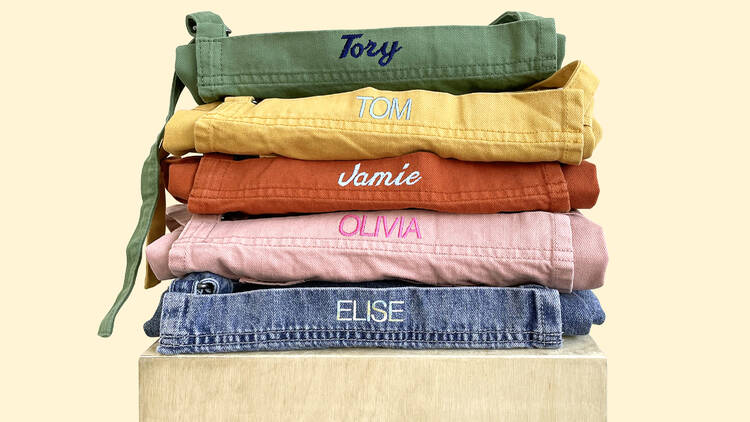 A stack of aprons in green, yellow, red, pink and denim with embroidered names sit folded on top of one another