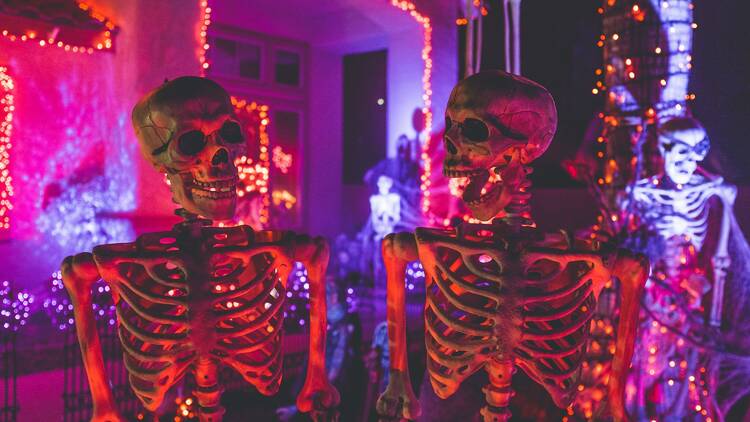 two skeletons near a white concrete building with red and purple fairy lights at night