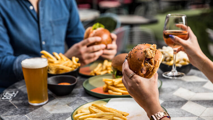 A person holds a burger and a glass of wine, with another person eating a burger and fries in the background