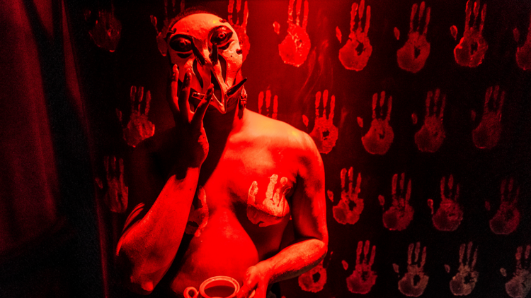 Performance artist Mara Maya Devi is bathed in red light and wears an alien like mask. The wall behind them is covered in red hand prints and they have hand prints on their chest.