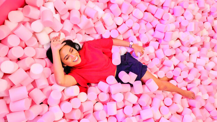 A woman in red shirt lays in a pool filled with giant pink marshmallows