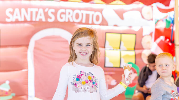 A smiling little girl holding an ice cream poses in front of a colourful backdrop that says 'Santa's Grotto'